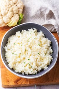 A gray bowl filled with cauliflower rice on top of the wooden cutting board. Part of a cauliflower head and part of a gray napkin are behind the bowl.