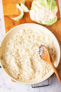 A wooden cutting board with a skillet on top filled with riced cauliflower. There is a head of cauliflower behind the skillet in the wooden spoon is in the skillet.