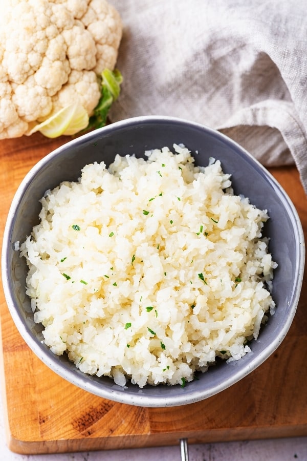 A gray bowl filled with cauliflower rice on top of the wooden cutting board. Part of a cauliflower head and part of a gray napkin are behind the bowl.