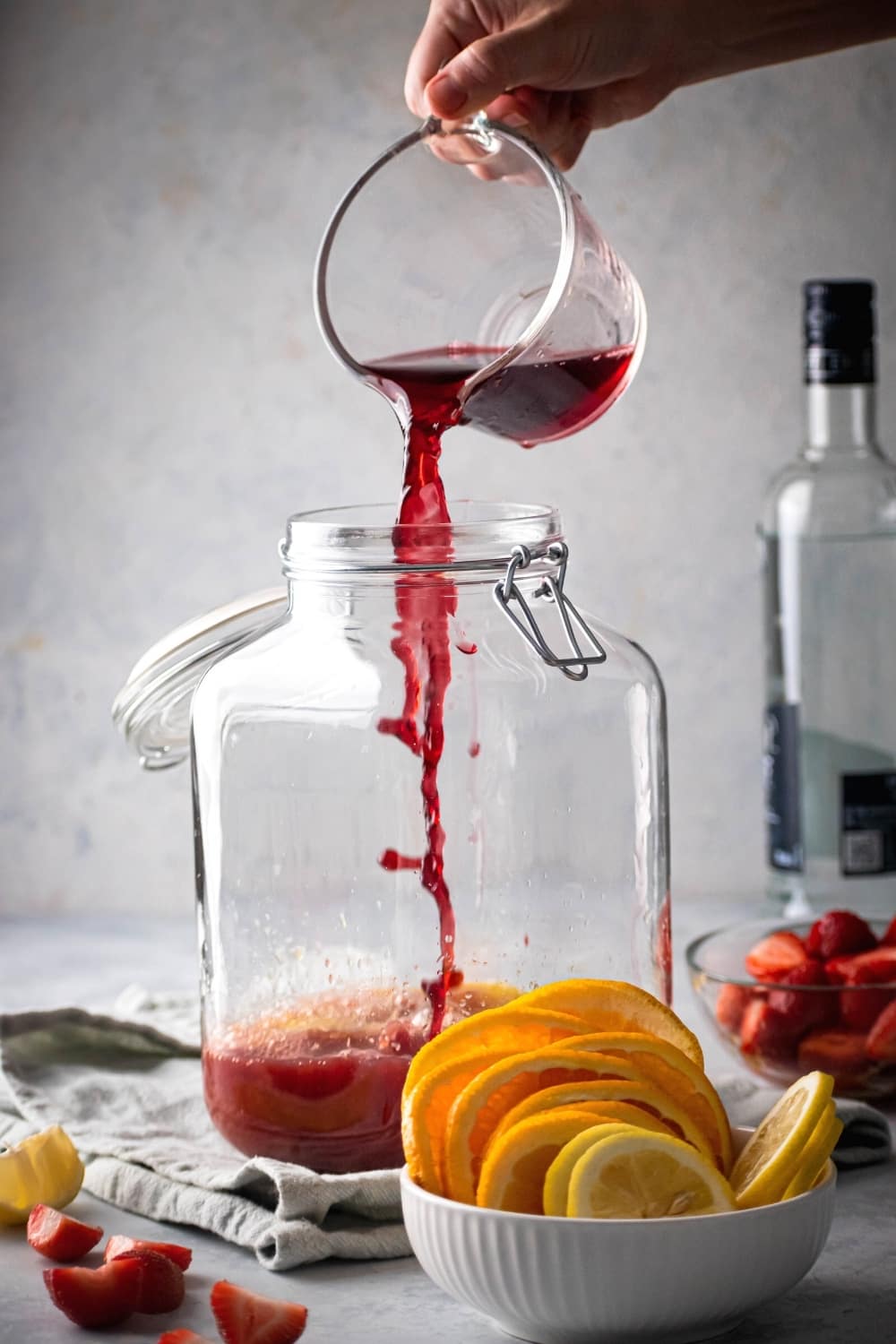 Glass gallon dispenser with some juice in it. I hand is pouring a cup of cranberry juice into it. Behind the jungle juice is a bottle of vodka in front of that a small glass bowl sliced strawberries and in front of that white bowl of orange slices.