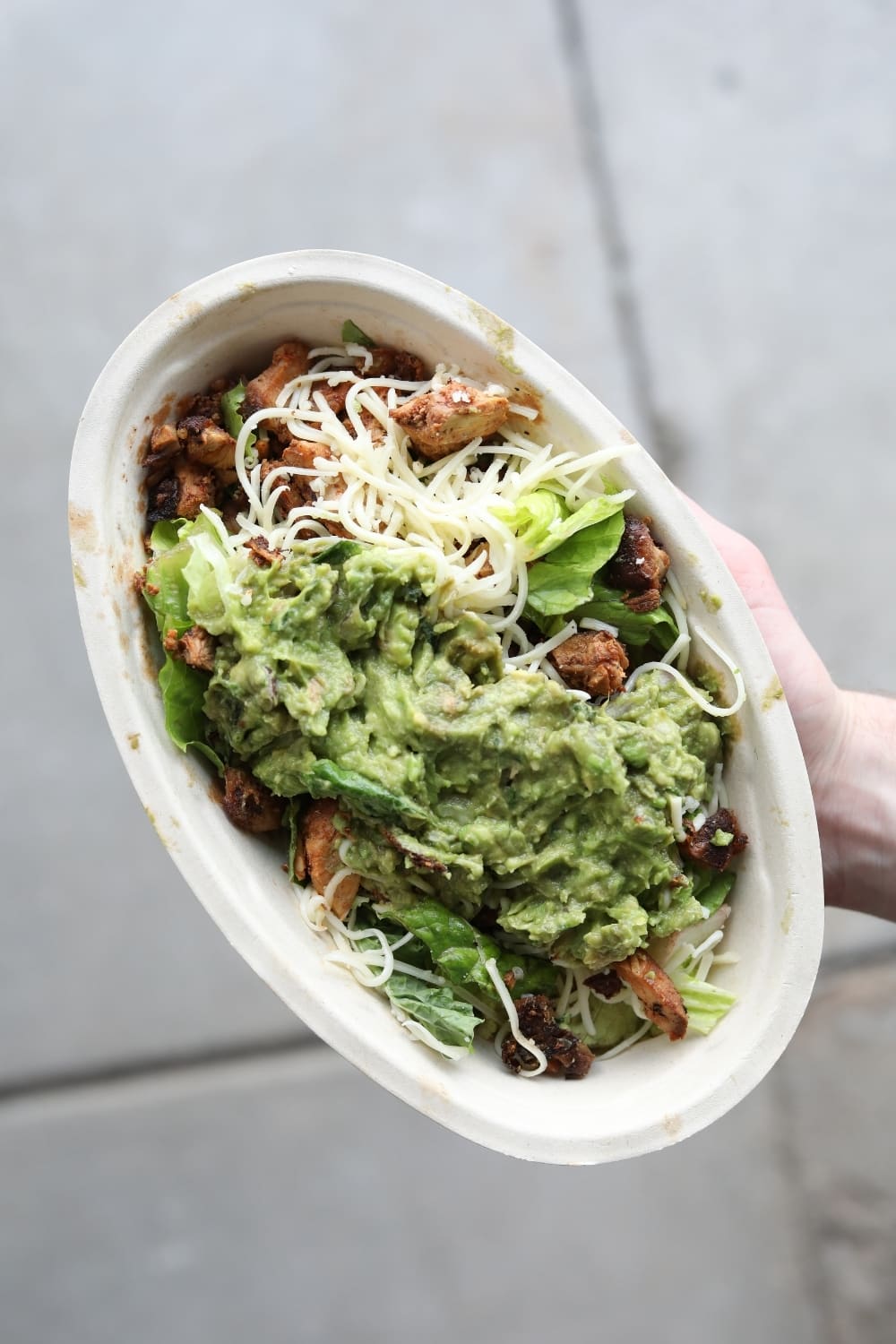 A hand holding a keto chipotle bowl with chicken, shredded cheese, lettuce, and guacamole.