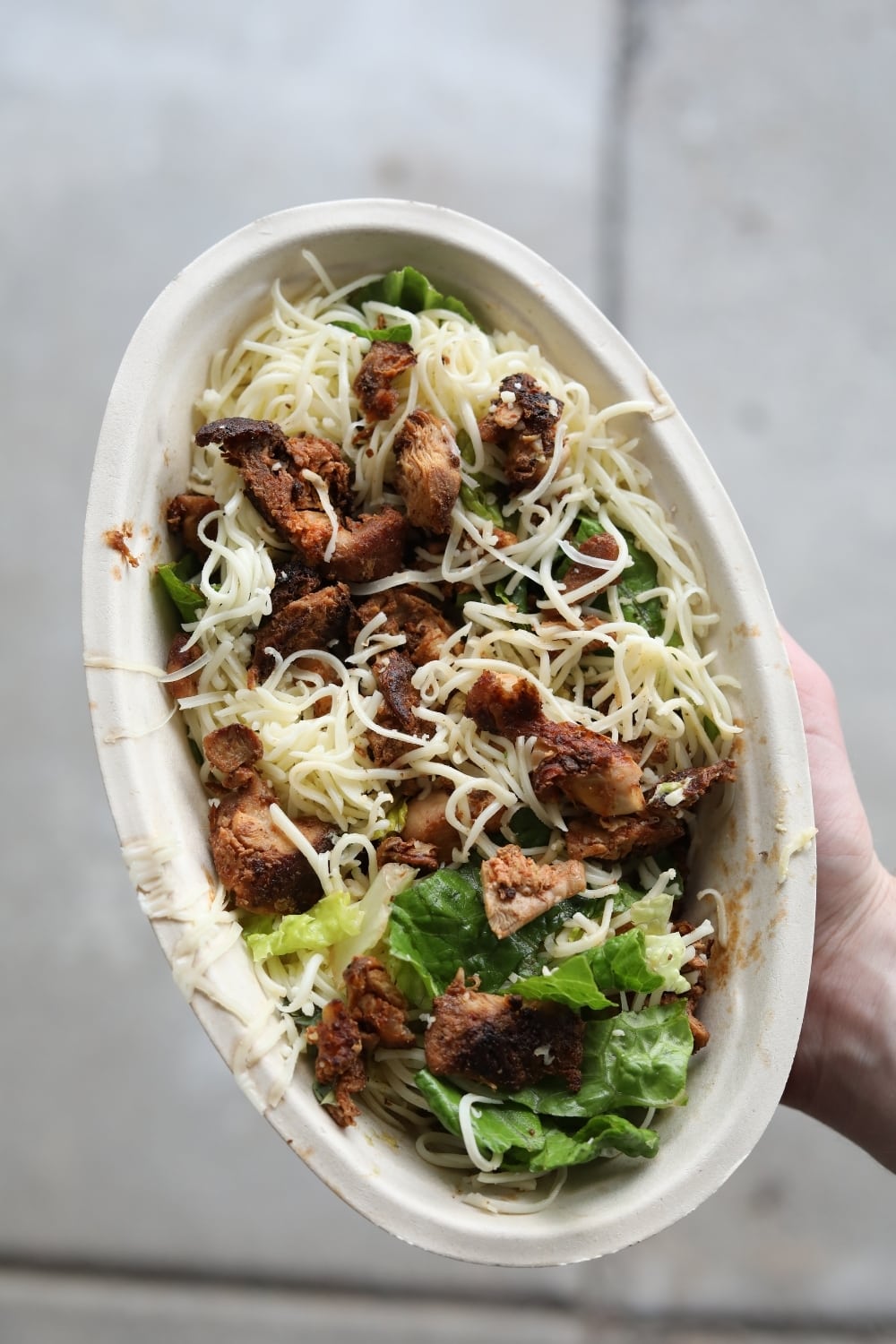 A hand holding a keto chipotle bowl with chicken, Romain lettuce, and shredded cheese.