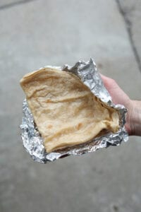 A hand holding a folded tortilla shell on a piece of tin foil.