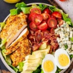 A white play with Cobb salad on it. Part of a glass jar filled with Dijon vinaigrette dressing is behind the plate and both are on a serving board.