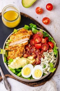 A white plate filled with a Cobb salad. The plate is on a serving tray and there is a glass jar of Dijon vinaigrette dressing behind it. A whole tomato and tomato sliced in half or next to the serving plate.
