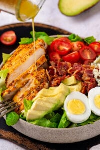 A white bowl filled with a Cobb salad. Part of a glass jar is drizzling Dijon vinaigrette dressing on the chicken portion of the salad.