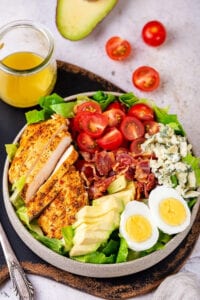 A white plate filled with a Cobb salad. Behind the plate is a glass jar with Dijon vinaigrette dressing in it and both of them are on a serving board. Sliced cherry tomato on whole cherry tomato or behind the serving board and part of an avocado is next to the tomatoes.