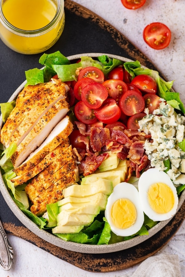 A white play with Cobb salad on it. Part of a glass jar filled with Dijon vinaigrette dressing is behind the plate and both are on a serving board.