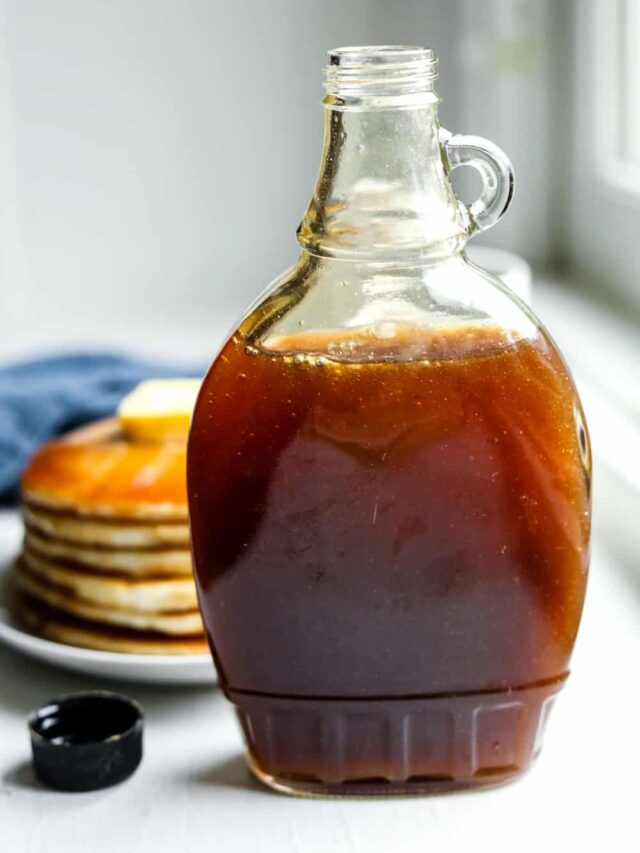 A bottle of maple syrup in front of a plate of pancakes.