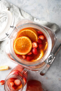 A glass gallon dispenser filled with jungle juice, orange slices, and sliced strawberries. In front of it is a a small glass jar filled with the same thing.