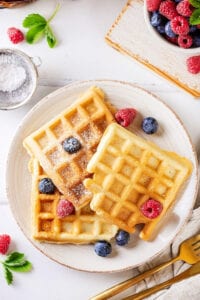 Three waffles on a white plate with a few blueberries and raspberries on top. Behind the plate is part of a bowl of blueberries and raspberries. In front of the plate is part of a golden fork and knife.
