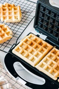 A waffle iron with a waffle and part of another waffle in it. There is a black wire rack next to it with one waffle and part of another waffle on it.