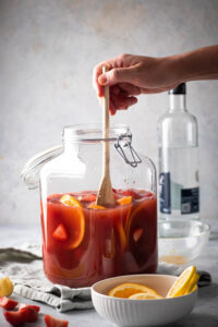 A glass dispenser filled with jungle juice, orange slices, and strawberry slices. Hand is stirring the jungle juice with a wooden spoon. In front of the jungle juice is a small white bowl with some orange slices and behind the jungle juice is a bottle of vodka.