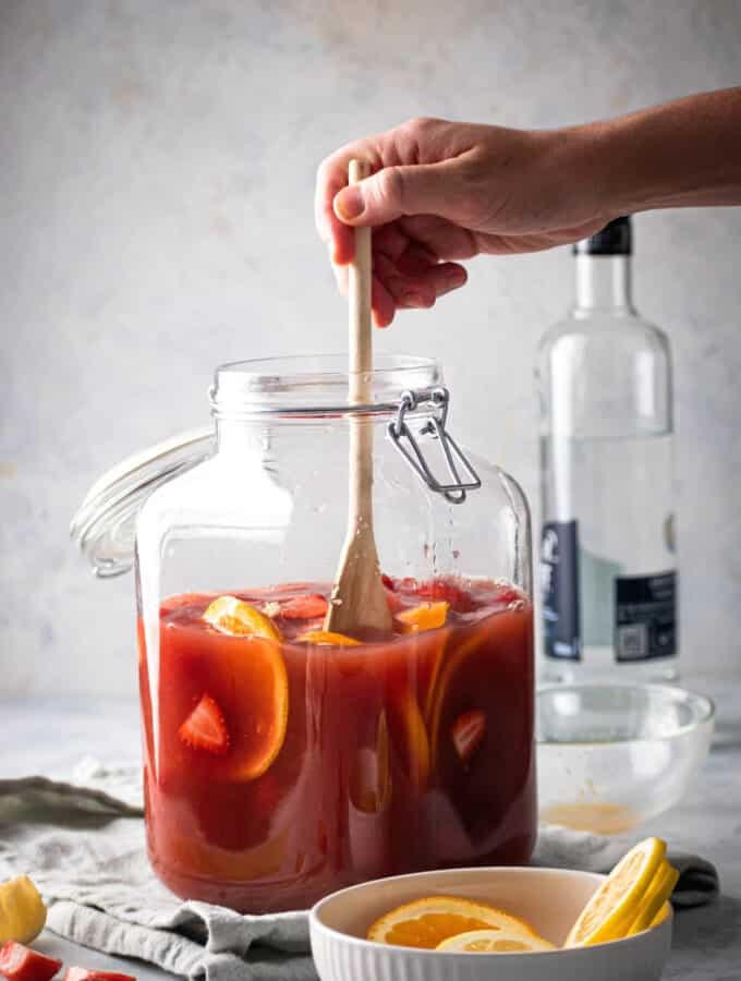 A glass dispenser filled with jungle juice, orange slices, and strawberry slices. Hand is stirring the jungle juice with a wooden spoon. In front of the jungle juice is a small white bowl with some orange slices and behind the jungle juice is a bottle of vodka.