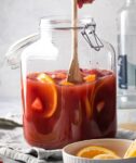 A glass dispenser filled with jungle juice, orange slices, and strawberry slices. Hand is stirring the jungle juice with a wooden spoon. In front of the jungle juice is a small white bowl with some orange slices and behind the jungle juice is a bottle of vodka.