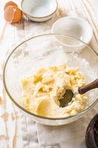A glass bowl filled with coconut macaroon batter with a spoon in it. There are two small bowls and a cracked egg shell behind it.