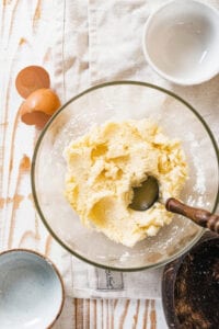 A glass bowl of coconut macaroon batter with a spoon in it. And egg shell is to the left of the bowl and two small empty bowls are on the sides of it.