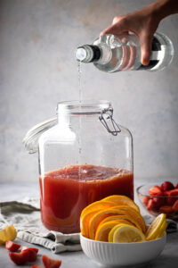 A glass gallon dispenser filled with juices. A hand is pouring vodka from a bottle into the juice. In front of the gallon dispenser is a small white bowl of sliced oranges in behind that small glass bowl of sliced strawberries.