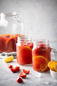 Two glass cups filled with jungle juice, strawberry slices, and orange slices, behind the glasses is half of a glass gallon jar filled with jungle juice. On the white counter everything is on there's a few slices of strawberries.
