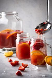 A glass jar filled with ice, an orange slice, and sliced strawberries. A little is pouring jungle juice into it and behind that glass is a filled glass of jungle juice. Behind the two glasses is half of a glass gallon jar filled with jungle juice. The glasses on the counter for a few sliced strawberries.