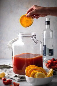 A glass gallon dispenser filled with jungle juice. In front of it is a small white bowl with orange slices and a hand is holding an orange slice, hovering over the jungle juice.