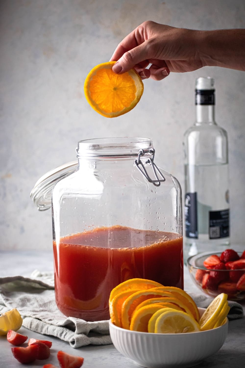 A glass gallon dispenser filled with jungle juice. In front of it is a small white bowl with orange slices and a hand is holding an orange slice, hovering over the jungle juice.