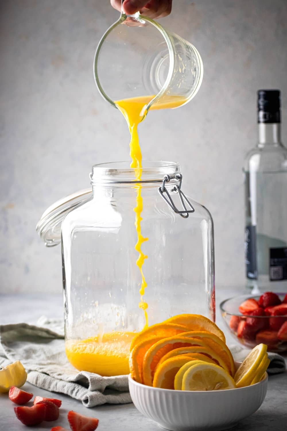 An empty glass gallon dispenser. A hand is pouring a cup of orange juice into it. In front of the dispenser is a small white bowl with orange slices and next to dispenser is a small glass bowl of sliced strawberries, behind the strawberries as a bottle of vodka.