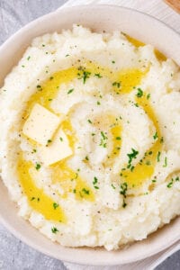 A white bowl filled with cauliflower mashed potatoes. There are two slices of butter with melted butter on the top. The warzone a white tablecloth on a gray counter.