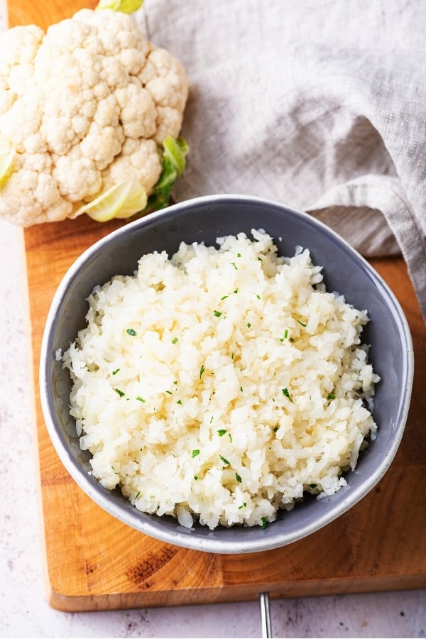 A grey bowl filled with cauliflower rice on a wooden cutting board. There's a head of cauliflower behind the bowl with a gray napkin next to it.