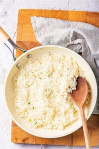 A wooden cutting board with a skillet filled with riced cauliflower. There is a wooden spoon in the skillet and a gray napkin behind the skillet.