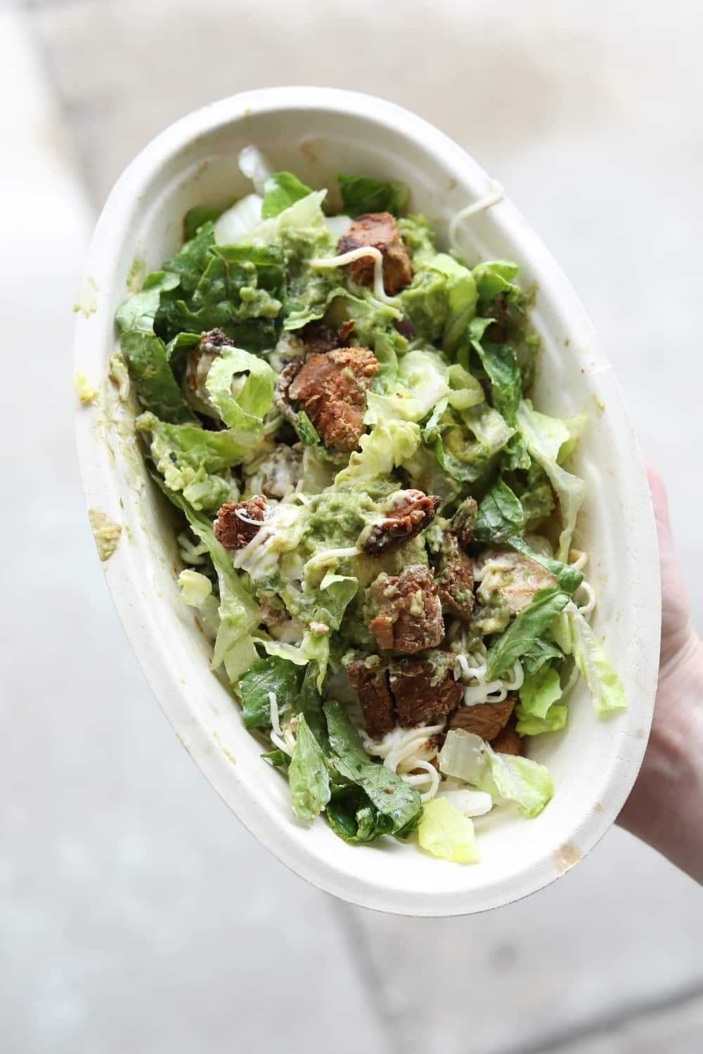 A hand holding a keto chipotle salad bowl with lettuce, steak, shredded cheese, and guacamole.