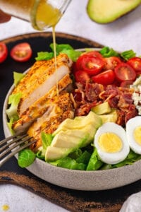 A white bowl filled with carbs salad. Part of a glass jar is drizzling Dijon vinaigrette dressing and chicken in the salad bowl. Four prongs are touching some of the pieces of chicken at the edge of the bowl.