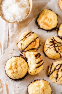 A couple of coconut macaroons on a piece of white parchment paper on a white tablecloth. Macaroons have chocolate drizzled on top and some chocolate on the bottom. To the back left of the macaroons is part of a small bowl with shredded coconut in it.