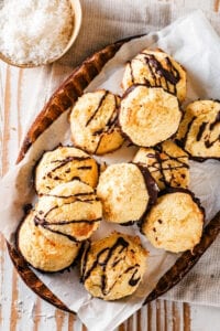 And oval bowl with white parchment paper in it with 11 coconut macaroons on top. Half of them have chocolate sauce drizzled on top and half of chocolate on the bottom of them. The bowl is on a gray tablecloth on a wooden table and behind tablecloth as part of a small bowl shredded coconut.