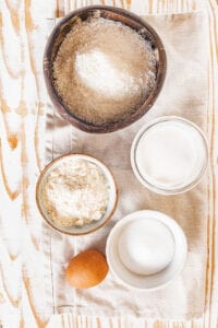 A medium sized bowl of finley shredded coconut, a small bowl of coconut flour in front of it, a small bowl of coconut milk to the right of it, and a small bowl of granulated erythritol and an egg. Everything is on a rectangular tablecloth on a wooden table.