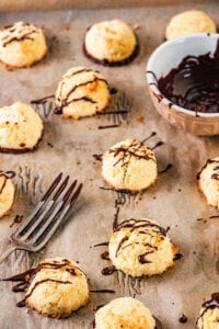 A piece of brown parchment paper with a bunch of coconut macaroons drizzled in chocolate sauce on it. There is a fork with chocolate on the prongs and part of a small bowl of chocolate sauce between the macaroons.