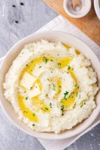 A white bowl filled with cauliflower mashed potatoes with two squares of butter and some melted butter on top. The balls on a white tablecloth on a gray counter and there is part of a wooden cutting board behind it.