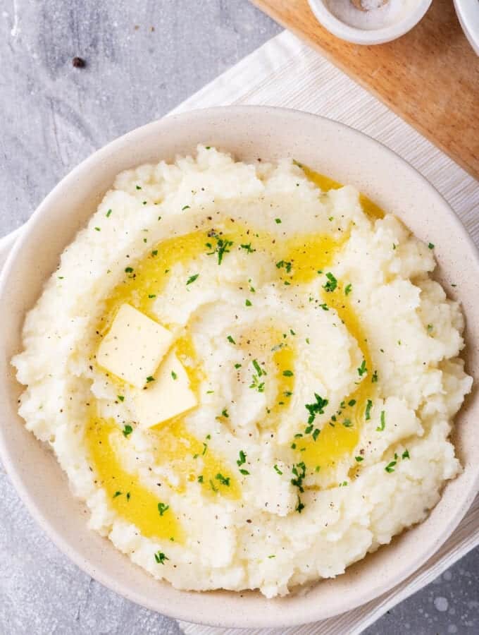 A white bowl filled with cauliflower mashed potatoes with two squares of butter and some melted butter on top. The balls on a white tablecloth on a gray counter and there is part of a wooden cutting board behind it.