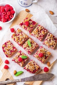 Three rows of three raspberry cheesecake bars on a piece of parchment paper on top of a wooden cutting board. There's a small bowl of raspberries behind the cutting board and a knife at the front of it.