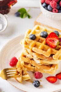 A stack of five waffles on a white plate with a few blueberries on the top waffle. The prongs of a fork is in front of the Stack with two pieces of waffle on it.
