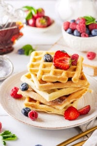 A white plate with a stack of five waffles on it. There are some sliced strawberries, blueberries, and raspberries on them. Behind the plate is a bowl filled with raspberries and blueberries.