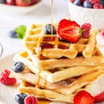 A stack of five waffles on a white plate. There is some maple syrup then drizzled over the waffles and a few raspberries, blueberries, and strawberries are on the waffles and plate.