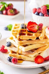 A stack of five waffles on a white plate. There is some maple syrup then drizzled over the waffles and a few raspberries, blueberries, and strawberries are on the waffles and plate.