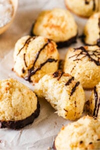 A couple of coconut macaroons on a piece of white parchment paper. One macaroon has a bite out of the front of it showing the inside.