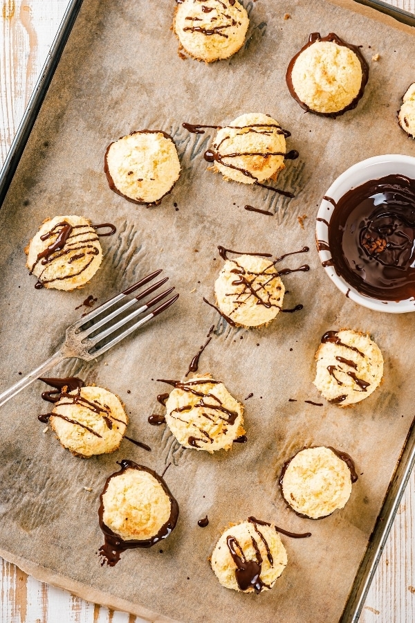 A baking sheet lined with brown parchment paper with a bunch of coconut macaroons on it. Some of the macaroons have chocolate sauce drizzled on top and some have chocolate on the bottom. There is a fork with chocolate on the prongs of it and part of a small bowl of chocolate sauce on the baking sheet.