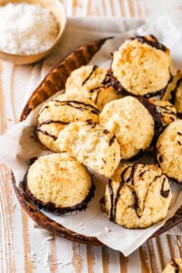 An oval brown bowl with a piece of parchment paper in it with a bunch of coconut macaroons on top. There is a bite out of one of the macaroons and several of them have chocolate sauce drizzled on top and others have chocolate on the bottom.