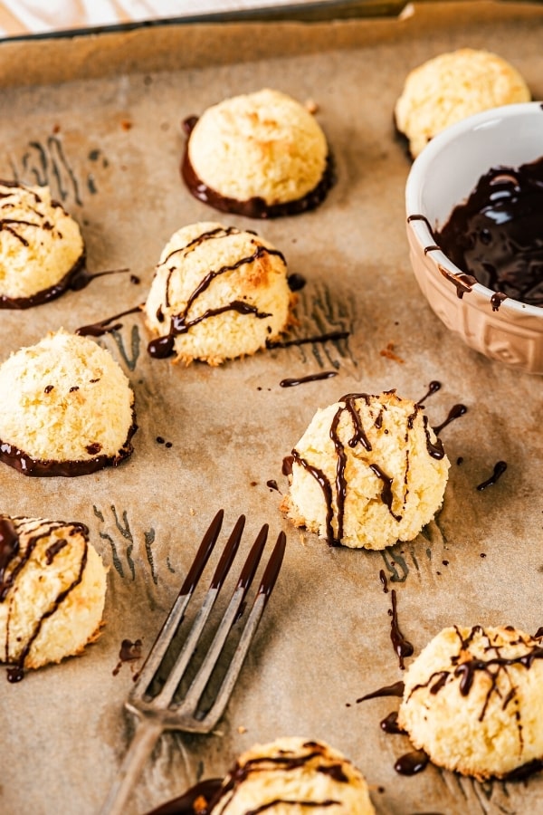 Several coconut macaroons with a few drizzled with chocolate and a few with chocolate on the bottom. All of the macaroons are on a piece of brown parchment paper on a baking tray and there is half of a small bowl of chocolate sauce and the head of a fork covered in chocolate around the macaroons.