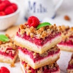 Three raspberry cheesecake bars stack on top of one another on a piece of white parchment paper. There are some raspberry cheesecake bars around the stack and part of a bowl of raspberries in the background.