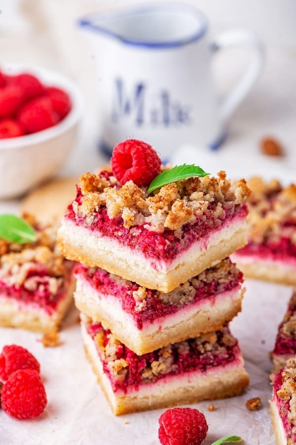 Three raspberry cheesecake bars stack on top of one another on a piece of white parchment paper. There are some raspberry cheesecake bars around the stack and part of a bowl of raspberries in the background.