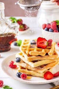 A stack of five waffles on a white plate. There is powdered sugar being sifted on the top of the waffles. There is part of a glass of maple syrup and a small bowl of blueberries and raspberries next to it.